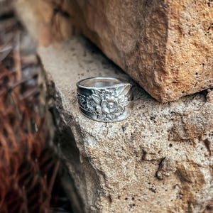 May include: A silver spoon ring with a floral design. The ring is sitting on a rock.