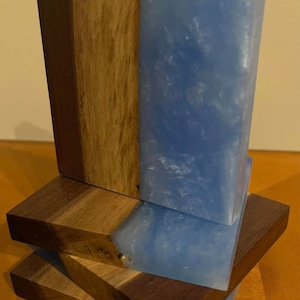 Puede incluir: Conjunto de cuatro posavasos hechos de madera y resina azul. Los posavasos tienen un acabado de madera natural y una resina azul claro con remolinos blancos. Los posavasos están apilados uno encima del otro.