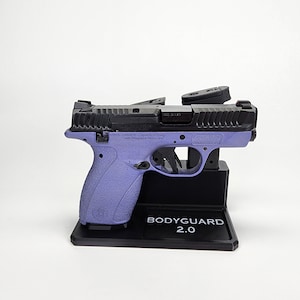 May include: A black and purple Smith & Wesson Bodyguard 2.0 handgun is displayed on a black stand. The gun has a black slide and a purple grip. The stand has the text "BODYGUARD 2.0".