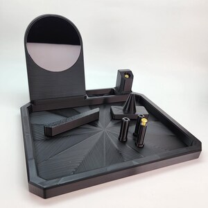 Pode incluir: Um organizador preto impresso em 3D com um pequeno espelho, um suporte c&ocirc;nico e quatro pequenos compartimentos cil&iacute;ndricos. O organizador tem uma superf&iacute;cie texturizada e um design geom&eacute;trico.