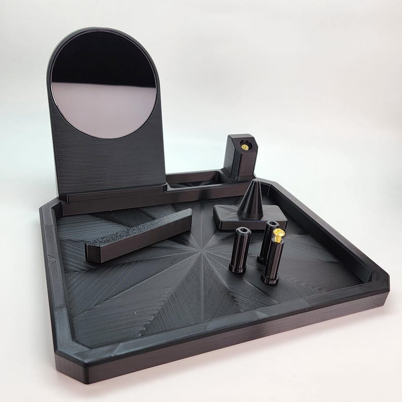 Pode incluir: Um organizador preto impresso em 3D com um pequeno espelho, um suporte c&ocirc;nico e quatro pequenos compartimentos cil&iacute;ndricos. O organizador tem uma superf&iacute;cie texturizada e um design geom&eacute;trico.