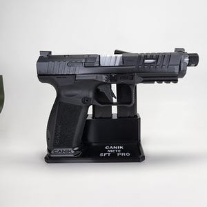 May include: A black Canik Mete SFT Pro handgun on a black stand. The gun has a textured grip and the Canik logo is visible. The stand has the text "CANIK METE SFT PRO". The gun is displayed against a white background.