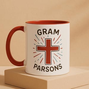 Peut inclure: Mug en céramique blanche avec intérieur et anse rouges. Le mug porte l'inscription "GRAM PARSONS" dans une police stylisée, avec une croix rouge au centre et des éclats rouges et bleus. Le mug est posé sur une surface beige.