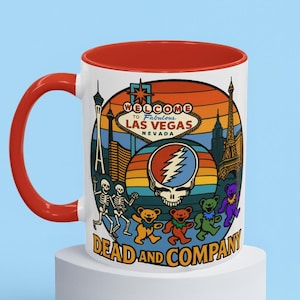以下が含まれることがあります： 赤色の内側とハンドルが付いた白いセラミックマグ。マグには、ラスベガスの看板、エッフェル塔、踊るクマのカラフルなグラフィックが描かれています。「Welcome to Fabulous Las Vegas Nevada」と「Dead and Company」の文字も表示されています。