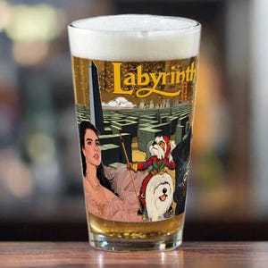 May include: A clear glass pint glass with a white foam head. The glass features a colorful graphic of a woman, a dog, and a creature from the movie Labyrinth. The text "Labyrinth" is printed on the glass.