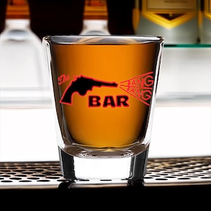 May include: A clear glass shot glass with a black silhouette of a gun and the text "The Bang Bar" in red.