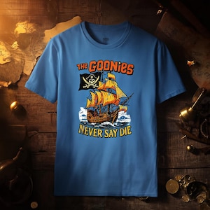 Puede incluir: Camiseta azul claro con un diseño de barco pirata y el texto "THE GOONIES NEVER SAY DIE". El barco tiene velas naranjas y una bandera con una calavera y huesos cruzados. La camiseta es de material suave.