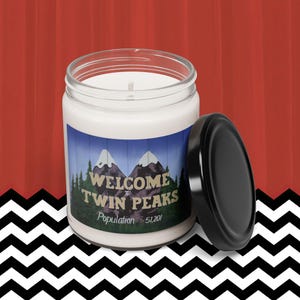 May include: A white candle with a black lid. The candle has a blue and white label that says "Welcome to Twin Peaks Population 51,201".