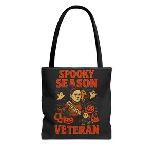 May include: Black tote bag with a Halloween-themed design. The design features the text "Spooky Season Veteran" in orange and white, along with illustrations of pumpkins, ghosts, and a character in a hat.