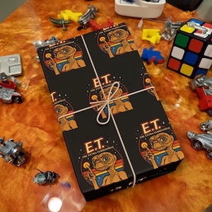 May include: A wrapped gift box featuring a black background with a repeating pattern of E.T. the Extra-Terrestrial. The design includes the character's image, the letters "E.T.", and the phrase "The Extra Terrestrial". The box is tied with twine.