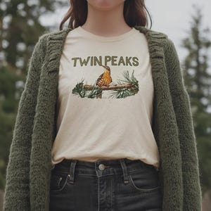 May include: Cream-colored t-shirt with the text "TWIN PEAKS" above a detailed illustration of a bird perched on a branch. The bird has brown and orange feathers. The shirt is worn with a green cardigan and black jeans.