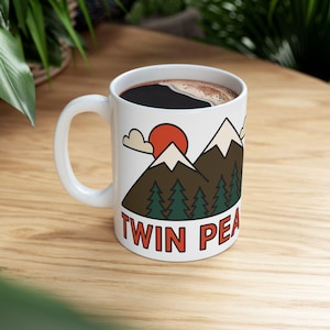 May include: White ceramic mug filled with dark coffee. The mug features a graphic of a mountain range with a red sun, green trees, and the text "TWIN PEA" in red letters.