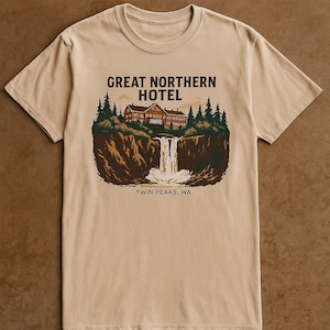 Op de afbeelding: Beige T-shirt met een afbeelding van het Great Northern Hotel, een waterval en de tekst "Twin Peaks, WA". Het ontwerp gebruikt bruin, groen en wit. Het shirt is gemaakt van een zacht, comfortabel materiaal.