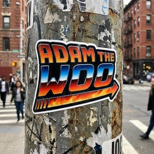 Peut inclure: Un autocollant sur un poteau métallique usé présente le texte "ADAM THE WOO" en orange et bleu, avec un motif de flamme et de flèche. L'autocollant est bordé de blanc et se détache sur une scène de rue urbaine floue.