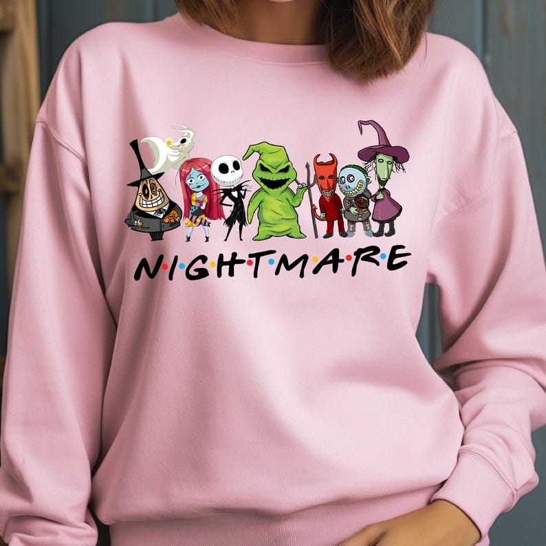 May include: A light pink sweatshirt featuring a graphic of characters from "The Nightmare Before Christmas." The word "NIGHTMARE" is printed across the front in colorful letters. The design includes Jack Skellington, Sally, and other characters.