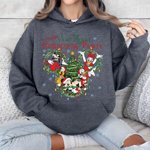 May include: Dark gray hoodie featuring a festive Christmas design. The graphic includes Mickey Mouse, Donald Duck, Goofy, and other Disney characters, along with a Christmas tree and the text "Mickey's Very Merry Christmas Party."