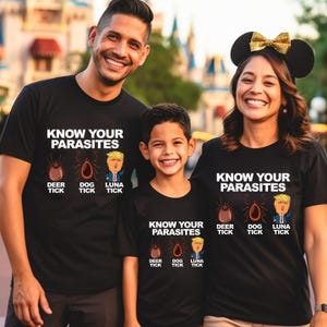 K&ouml;nnte beinhalten: Schwarze T-Shirts mit dem Text "KNOW YOUR PARASITES" und Illustrationen einer Hirschzecke, einer Hundezecke und einer Cartoonfigur. Die Shirts werden von einer dreik&ouml;pfigen Familie getragen.