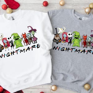 May include: Two sweatshirts, one white and one gray, each featuring a graphic of characters from "The Nightmare Before Christmas." The word "NIGHTMARE" is printed in a colorful font below the characters. Christmas ornaments are scattered around.