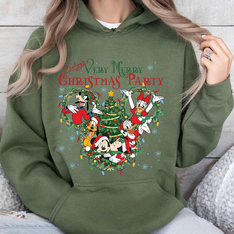 May include: Olive green hoodie featuring a festive design. The graphic includes Mickey Mouse, Donald Duck, Goofy, and Daisy Duck, surrounding a Christmas tree. The text "Mickey's Very Merry Christmas Party" is printed above the characters.
