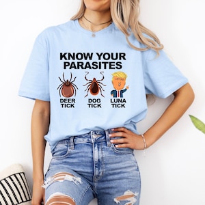 K&ouml;nnte beinhalten: Hellblaues T-Shirt mit dem Text "KNOW YOUR PARASITES" &uuml;ber Illustrationen einer Hirschzecke, einer Hundezecke und einer Cartoonfigur. Das T-Shirt wird von einer Person in Jeans getragen.