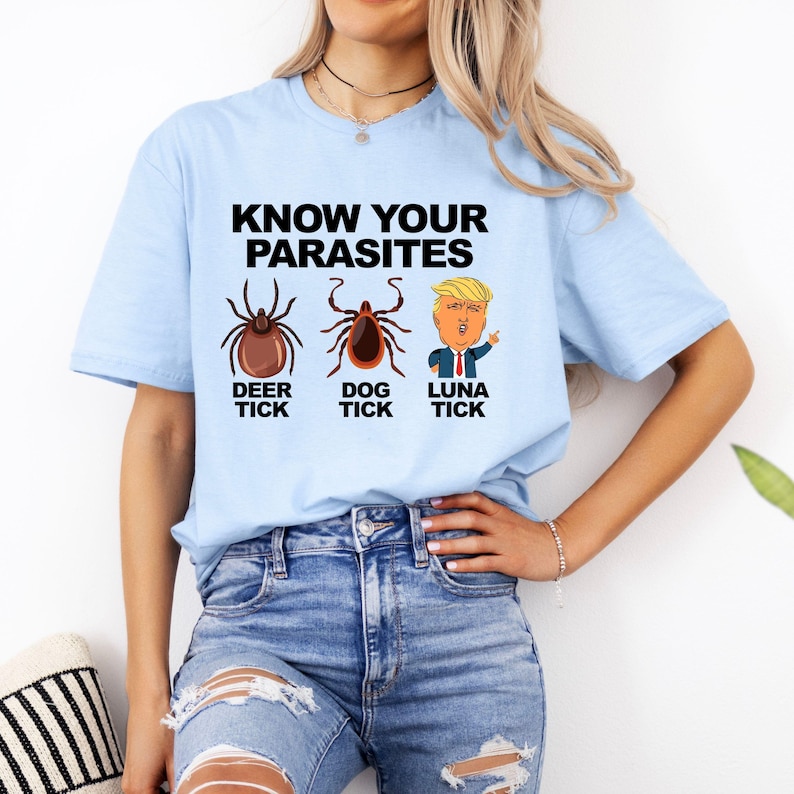 K&ouml;nnte beinhalten: Hellblaues T-Shirt mit dem Text "KNOW YOUR PARASITES" &uuml;ber Illustrationen einer Hirschzecke, einer Hundezecke und einer Cartoonfigur. Das T-Shirt wird von einer Person in Jeans getragen.