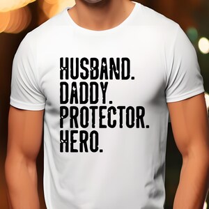 May include: White t-shirt with the words "HUSBAND. DADDY. PROTECTOR. HERO." printed in black, bold letters. The shirt is a crew neck style and appears to be made of a soft material.