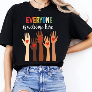 May include: Black t-shirt with the text "EVERYONE is welcome here" in rainbow colors. Five hands of different skin tones are raised with red hearts in their palms.