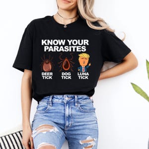 K&ouml;nnte beinhalten: Schwarzes T-Shirt mit dem Text "KNOW YOUR PARASITES" &uuml;ber Illustrationen einer Hirschzecke, einer Hundezecke und einer Cartoon-Figur. Das T-Shirt wird von einer Person getragen, die eine Jeans tr&auml;gt.