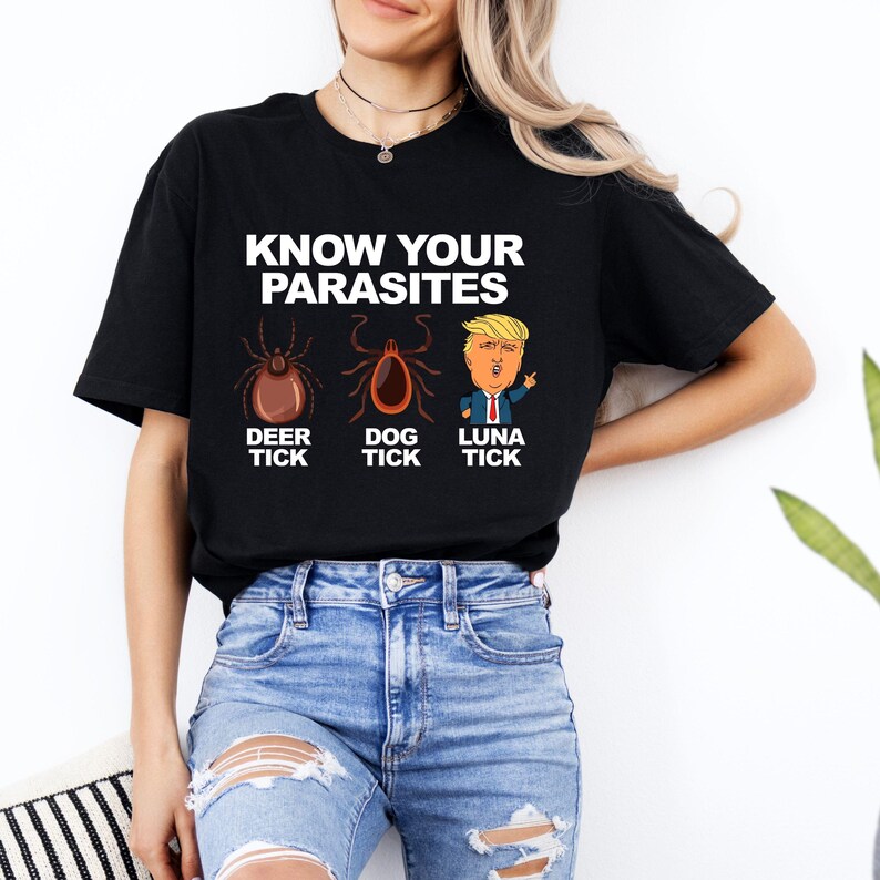 K&ouml;nnte beinhalten: Schwarzes T-Shirt mit dem Text "KNOW YOUR PARASITES" &uuml;ber Illustrationen einer Hirschzecke, einer Hundezecke und einer Cartoon-Figur. Das T-Shirt wird von einer Person getragen, die eine Jeans tr&auml;gt.