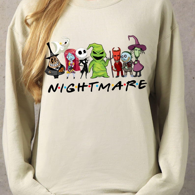 May include: A beige sweatshirt featuring a graphic of characters from "The Nightmare Before Christmas" with the word "NIGHTMARE" in a colorful font. The design includes Jack Skellington, Sally, and other spooky figures.