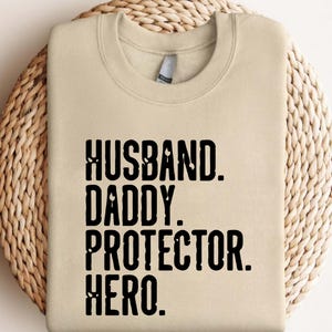 May include: A beige sweatshirt with the words "HUSBAND. DADDY. PROTECTOR. HERO." printed in bold black letters. The sweatshirt is displayed on a woven, circular surface.