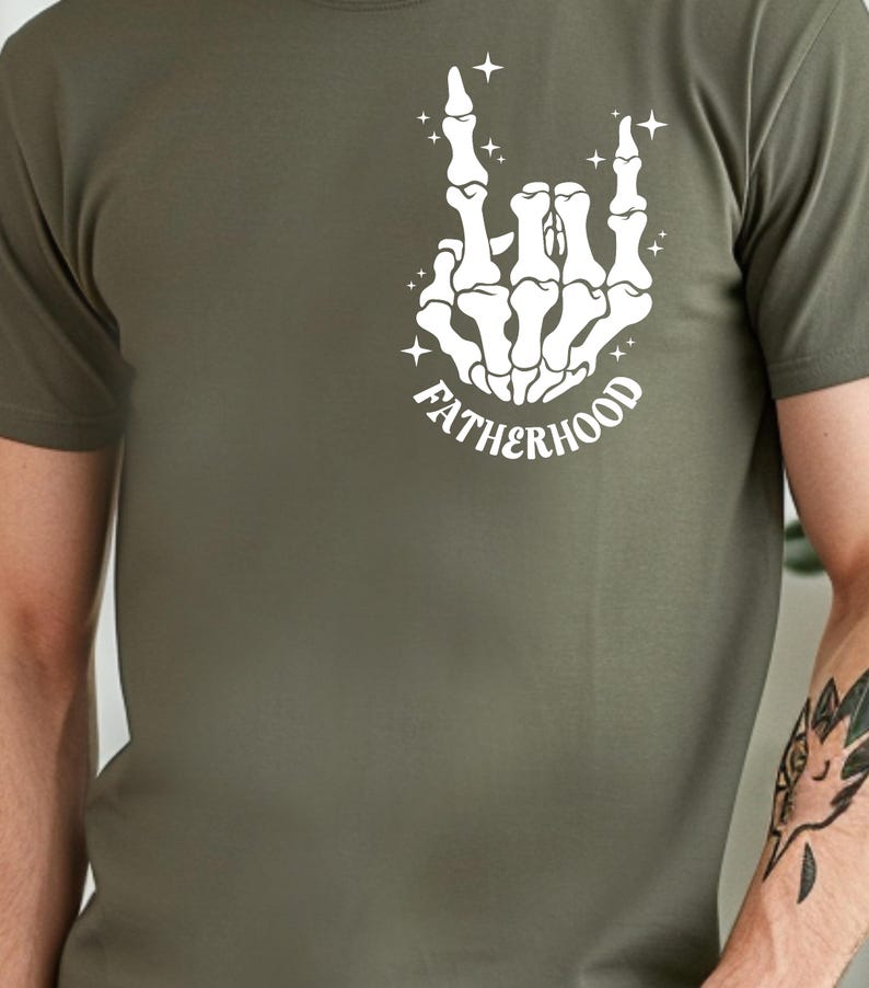 K&ouml;nnte beinhalten: Olivgr&uuml;nes T-Shirt mit einer wei&szlig;en Skeletthand, die eine Rock'n'Roll-Geste macht. Das Wort "FATHERHOOD" ist unter der Hand gebogen. Das Design enth&auml;lt Sternakzente.