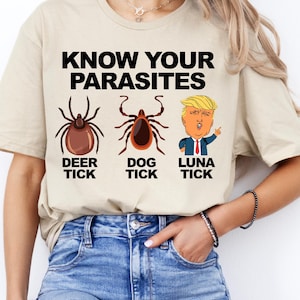 K&ouml;nnte beinhalten: Beiges T-Shirt mit dem Text "KNOW YOUR PARASITES" &uuml;ber Illustrationen einer Hirschzecke, einer Hundezecke und einer Karikatur einer Person mit dem Text "LUNA TICK". Das Shirt wird von einer Person getragen, die Jeans tr&auml;gt.