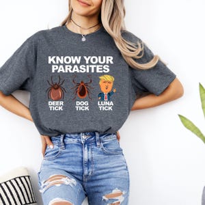 K&ouml;nnte beinhalten: Ein dunkelgraues T-Shirt mit dem Text "KNOW YOUR PARASITES" &uuml;ber Illustrationen einer Hirschzecke, einer Hundezecke und einer Karikatur einer Person. Das T-Shirt wird von einer Person getragen, die eine blaue Jeans tr&auml;gt.