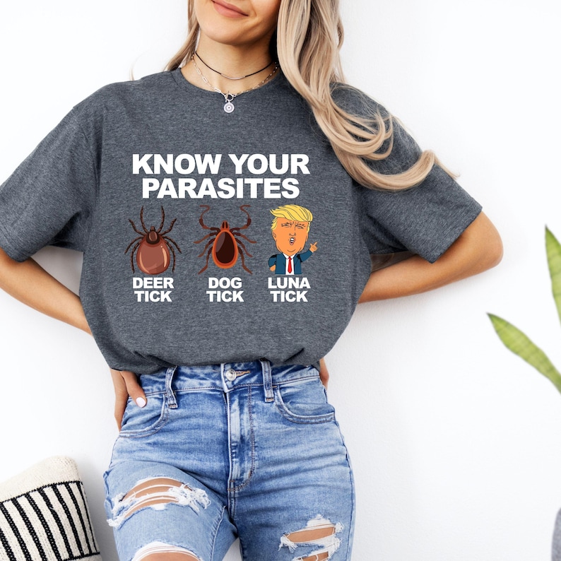 K&ouml;nnte beinhalten: Ein dunkelgraues T-Shirt mit dem Text "KNOW YOUR PARASITES" &uuml;ber Illustrationen einer Hirschzecke, einer Hundezecke und einer Karikatur einer Person. Das T-Shirt wird von einer Person getragen, die eine blaue Jeans tr&auml;gt.