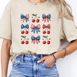May include: Beige t-shirt featuring a grid of patriotic bows and cherries. The bows are red, white, and blue, with star and stripe patterns. The cherries are red with green stems. A casual, festive design.