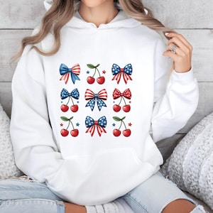 May include: White hoodie featuring a patriotic design of bows and cherries. The bows are in red, white, and blue, with star and stripe patterns. The cherries are red with green stems. A casual and festive top.