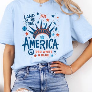 May include: Light blue t-shirt with a patriotic design. The shirt features the text "LAND OF THE FREE" and "AMERICA" in navy blue, red, and white. The design includes stars, a peace sign, and the year "1776".