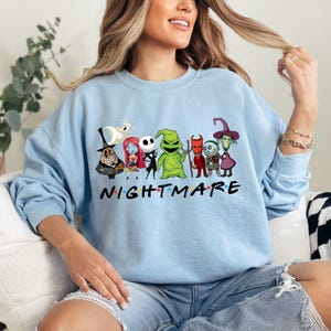 May include: Light blue sweatshirt featuring a graphic of characters from "The Nightmare Before Christmas" with the word "NIGHTMARE" in a colorful font. The design includes Jack Skellington, Sally, and Oogie Boogie.