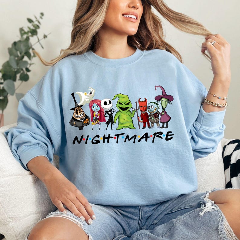 May include: Light blue sweatshirt featuring a graphic of characters from "The Nightmare Before Christmas" with the word "NIGHTMARE" in a colorful font. The design includes Jack Skellington, Sally, and Oogie Boogie.