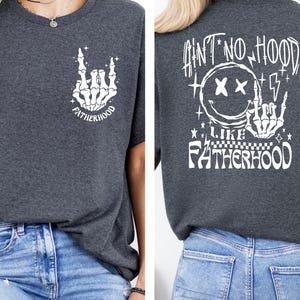 K&ouml;nnte beinhalten: Dunkelgraues T-Shirt mit wei&szlig;en Grafikdesigns. Die Vorderseite zeigt eine Skeletthand in einer Rock-and-Roll-Geste mit dem Wort "FATHERHOOD". Auf der R&uuml;ckseite befindet sich ein Smiley mit durchgestrichenen Augen, Blitzen und dem Satz "AIN'T NO HOOD LIKE FATHERHOOD".