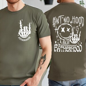 K&ouml;nnte beinhalten: Olivgr&uuml;nes T-Shirt mit einer wei&szlig;en Skeletthand-Grafik, die eine Rock'n'Roll-Handbewegung ausf&uuml;hrt, mit dem Wort "FATHERHOOD" darunter. Auf der R&uuml;ckseite des Shirts steht der Text "AIN'T NO HOOD LIKE FATHERHOOD" mit einem Smiley und Blitzen.