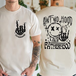 K&ouml;nnte beinhalten: Cremefarbene T-Shirts mit schwarzen Grafikdesigns. Eines zeigt eine Skeletthand mit dem Wort "FATHERHOOD". Das andere hat ein Smiley, einen Blitz und den Text "AIN'T NO HOOD LIKE FATHERHOOD".