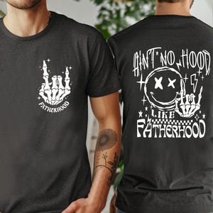 K&ouml;nnte beinhalten: Zwei dunkelgraue T-Shirts mit wei&szlig;en Grafikdesigns. Ein Shirt zeigt eine Skeletthand mit dem Wort "FATHERHOOD". Das andere Shirt hat den Text "AIN'T NO HOOD LIKE FATHERHOOD" mit einem Smiley und einer Skeletthand.