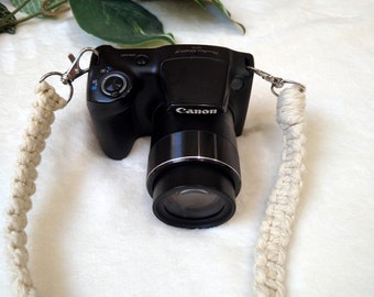 May include: A black Canon digital camera with a white braided strap. The camera has a zoom lens and a silver button on the top.