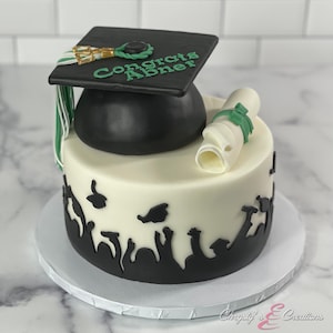 May include: A two-tiered white cake decorated with a black graduation cap, a green and white diploma, and black silhouettes of people throwing graduation caps in the air. The cap has the text "Congrats Abner" on it.