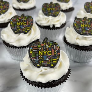 May include: A close-up of several chocolate cupcakes decorated with white frosting and black apple-shaped toppers with colorful NYC themed designs. The toppers feature the words "NYC", "WORK", "MEET", "LOVE", "PLAY", "HAPPY", "NOW", "HERE", and "PRAY".