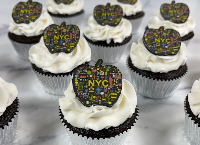 May include: A close-up of several chocolate cupcakes decorated with white frosting and black apple-shaped toppers with colorful NYC themed designs. The toppers feature the words "NYC", "WORK", "MEET", "LOVE", "PLAY", "HAPPY", "NOW", "HERE", and "PRAY".