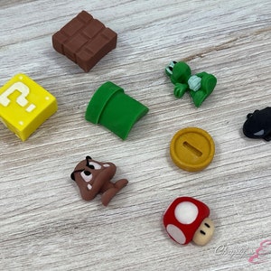 May include: A collection of colorful, miniature figurines inspired by the Super Mario video game series. Includes a brown brick, a yellow question mark block, a green pipe, a green turtle, a black cannon, a gold coin, a brown Goomba, and a red mushroom.