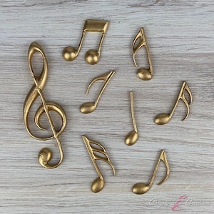 May include: Gold-colored musical notes and a treble clef are arranged on a light wood-grain surface. The collection includes various note types, such as eighth notes and quarter notes, all in a metallic gold finish. The image showcases musical decor.